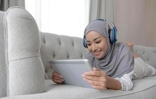 Relaxed and happy asian Muslim woman with hijab laying on sofa, listening music on headphones and using digital tablet. photo