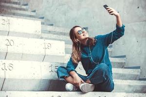 foto de una alegre y linda joven estudiante sonriente con gafas de sol al aire libre usando un teléfono móvil charlando escuchando música con auriculares.