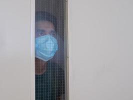 Lonely man in medical mask looking through the window. Isolation at home for self quarantine. Concept home quarantine, prevention COVID-19. Coronavirus outbreak situation photo