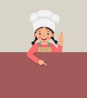 cute little girl in chef hat and apron standing behind empty board showing thumb up pointing finger to blank banner for copy space vector