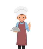 lindo niñito con sombrero de chef y delantal sosteniendo una bandeja mostrando el pulgar hacia arriba vector