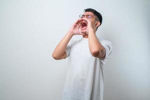 joven y apuesto hombre asiático con camisa informal y estilo de gafas gritando y gritando fuerte a un lado con la mano en la boca. concepto de comunicación foto