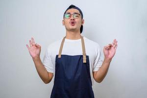 Young handsome asian barista man wearing apron standing relax and smiling with eyes closed doing meditation gesture with fingers. Yoga concept. photo