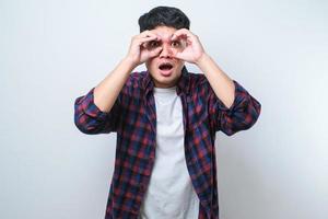 Young handsome man wearing casual shirt doing ok gesture like binoculars sticking tongue out, eyes looking through fingers. photo