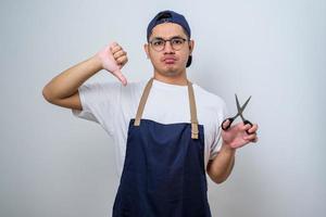 Young Asian barista man wearing apron holding scissors with unhappy face, negative sign showing dislike with thumbs down, rejection concept photo