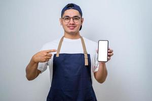 Young handsome asian barista man wearing apron pointing finger at mobile screen, showing app and smiling photo