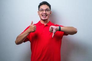 Young handsome Asian man wearing casual shirt and glasses  smilling, doing thumbs up and down, disagreement and agreement expression photo