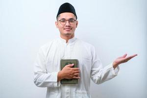 Asian muslim man smiling holding al quran book in hands and showing something on his side photo