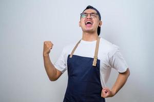 Asian barista man wearing apron standing very happy and excited doing winner gesture with arms raised, smiling and screaming for success. Celebration concept. photo
