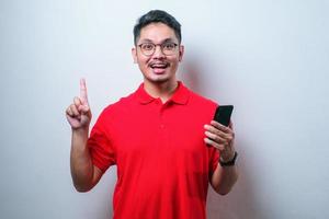 Asian young man showing happy expression with finger pointing up while holding mobile phone photo
