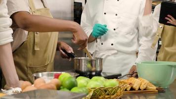 primo piano del corso di cucina per hobby, lo chef maschio senior in uniforme da cuoco insegna ai giovani studenti delle lezioni di cucina a mescolare e mescolare gli ingredienti per i pasticcini, le torte di frutta nella cucina in acciaio inossidabile del ristorante. video