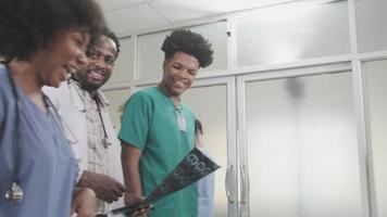 grupo de praticantes, médico afro-americano profissional com equipe de jovens estudantes de medicina, caminhada feliz e discutir filme de raio-x de diagnóstico no exame ambulatorial de saúde no hospital. video