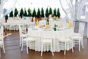 Round dinner tables covered with blue cloth stand in a white wedding pavilion photo
