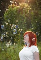 Young woman in a white T-shirt is playing with soap bubbles. Happy red-haired girl in the sun. photo