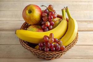 Ripe fruits in wicker basket on wooden background. Bananas, apples and grapes. Harvest concept. photo