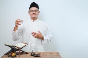hombre musulmán asiático sonriendo y pulgares hacia arriba mientras va a beber un vaso de agua para romper el ayuno foto