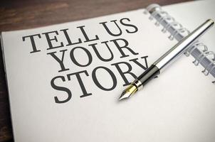 Text TELL US YOUR STORY on paper notepad and pen on wooden background photo
