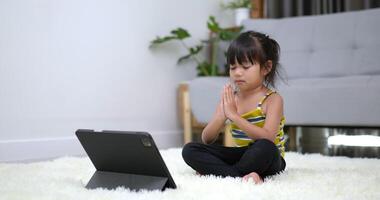 Slow-motion shot, klein Aziatisch meisje zittend op tapijt sloot haar oog en plaats twee handen op de borst in meditatie asana pose training op tablet in de woonkamer thuis video