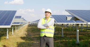 Dolly shot, portrait Asian young engineer wear white helmet standing and crossing arms with smile at solar farm, solar panel in background video