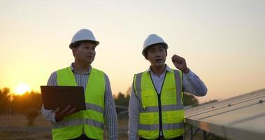 Two Asian young engineers walking along rows of photovoltaic panels in solar farm, They use laptop computer and talking together