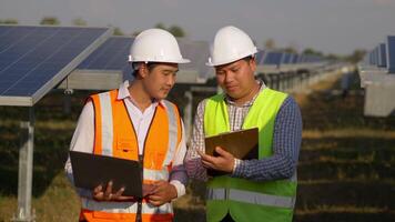 un ingeniero inspector asiático sostiene un tablero de control y un joven especialista usa una computadora portátil, dos ingenieros discuten y se alegran del éxito durante el trabajo en una granja solar, un panel solar en segundo plano video