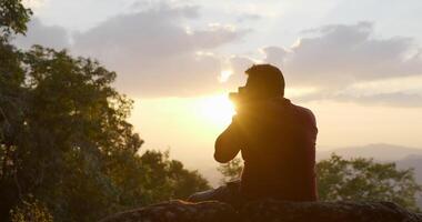 Slow motion in backlit shot, Young backpacker man sitting on rocky cliffs use digital camera take a photo while s in forest video