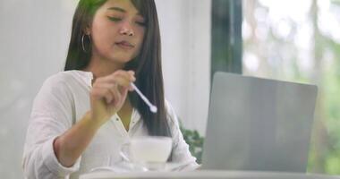 primo piano, bella donna d'affari asiatica che beve caffè mentre è seduto e lavora al computer portatile nel suo posto di lavoro. concetto di business e stile di vita. video