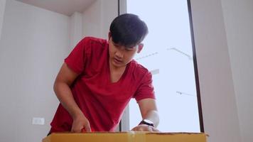 tiro em câmera lenta, jovem asiático usa fita adesiva para fechar a caixa de papelão, selando a caixa grande para mover, novo conceito de casa, foco seletivo
