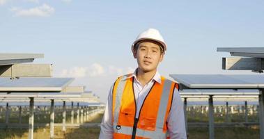 Dolly shot, portrait Asian young engineer wear white helmet standing and crossing arms with smile at solar farm, solar panel in background