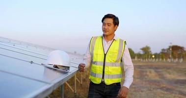 Asian young engineer man wearing white helmet and smile before start works in solar farm, solar panels on background