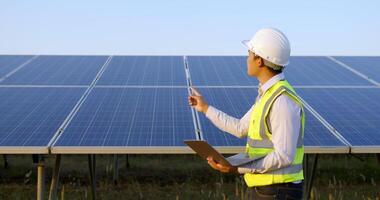 Young Asian technician man walking to checking operation of sun and photovoltaic solar panel and typing on laptop computer while working in solar farm video