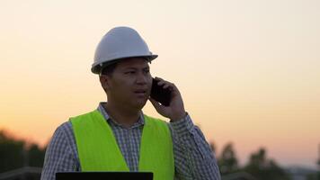 Young Asian Technician Engineer man hold laptop computer and upset during talking on smartphone while check efficiency of sun for examination on solar panel construction on sunset video