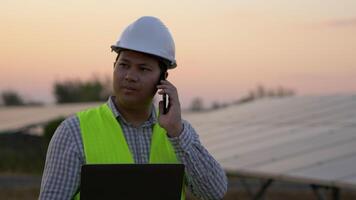 jonge aziatische technicus ingenieur man gebruikt laptopcomputer en praat met smartphone terwijl hij de efficiëntie van de zon controleert voor onderzoek op de constructie van zonnepanelen bij zonsondergang video