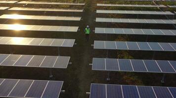 Aerial drone view, Flight over solar panel farm and young engineer wearing white helmet talking on smartphone and walking between solar panels row on sunset video