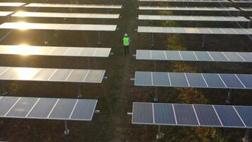 toma en cámara lenta, vista aérea de drones, vuelo sobre la granja de paneles solares, espalda de un joven ingeniero hablando por teléfono inteligente y caminando entre la fila de paneles solares al atardecer video