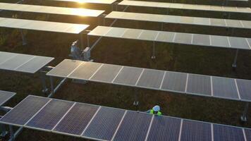 drone vanuit de lucht, vlucht over zonnepaneelboerderij, jonge ingenieur met helm die de werking van de zon controleert en de netheid van fotovoltaïsche zonnepanelen bij zonsondergang video