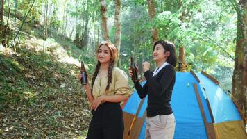 Two beautiful Asian women friends travelers relaxing with dance and cheering and drinking beer during camping in nature park, laugh and smile with happy together video