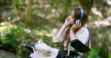 jovem mulher asiática sente-se em uma cadeira perto do córrego, ouvindo música do tablet com fones de ouvido sem fio alegremente enquanto acampa na floresta