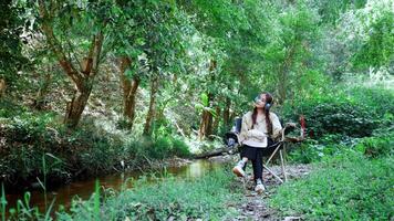 Young asian woman wearing hat sit in a chair near the stream, listening to music from headphones happily while camping in the woods video