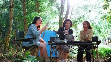 Young asian woman cooking and her friend enjoy to use smartphone take a photo the meal in pot, They are talk and laugh with fun together while camping in nature park