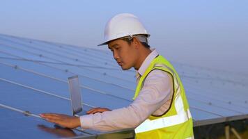 Young Asian technician man typing on laptop computer during checking operation of sun and photovoltaic solar panel while working in solar farm video