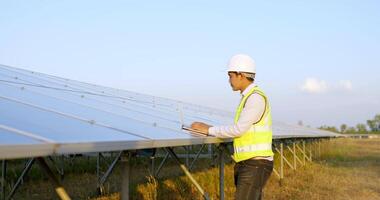 Young Asian technician man checking operation of sun and photovoltaic solar panel and typing on laptop computer while working in solar farm video