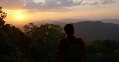 colpo retroilluminato, vista posteriore ravvicinata giovane escursionista che cammina, alza le mani e si guarda intorno con felice sulla cima della montagna rocciosa al tramonto video