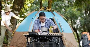 Asian man preparing pitch a tent in camping while the other man was preparing food. in camping. Cooking set front ground. Outdoor cooking, traveling, camping, lifestyle concept. video