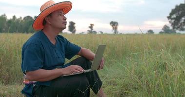 Portrait of Asian farmer man in hat with loincloth checking the growth of rice fields on laptop while sitting at the paddy field.