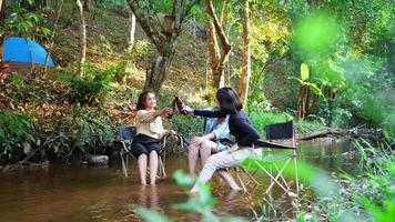 prise de vue à la main, un groupe de jeunes femmes asiatiques boivent de la bière sur leurs chaises et trempent leurs pieds dans le ruisseau en campant dans le parc naturel, elles aiment parler et rire ensemble. video