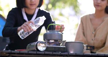Handaufnahme, Nahaufnahme der Hand einer jungen Frau, die Wasser aus der Flasche in den Wasserkocher goss, um mit ihren Freundinnen vor dem Campingzelt im Wald heißen Kaffee zu trinken video
