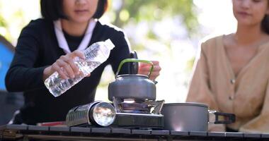 colpo a mano, mano ravvicinata della giovane donna versò l'acqua dalla bottiglia nel bollitore per far bollire per bere un caffè caldo con le sue amiche davanti alla tenda da campeggio nella foresta video