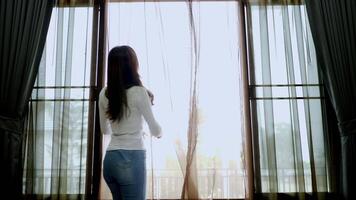 vista trasera de una joven con camisa blanca y jeans caminando para abrir la cortina y de pie mirando por la ventana para disfrutar con un hermoso cielo al aire libre video