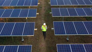 Aerial drone view, Flight over solar panel farm and young engineer wearing white helmet talking on smartphone and walking between solar panels row video